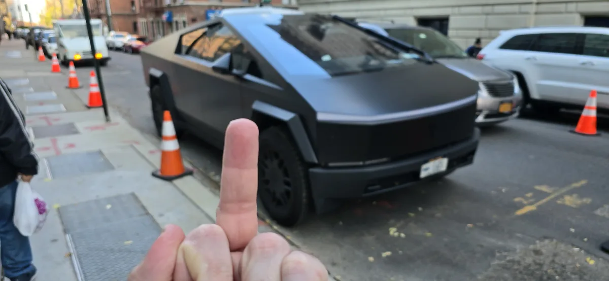 A close-up of a person's hand giving the middle finger, with a black Tesla Cybertruck parked on a city street in the…