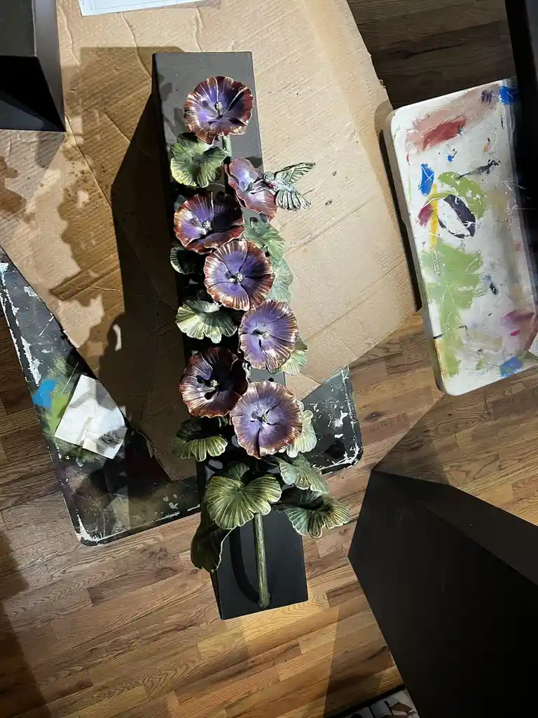 A top-down photo shows a handmade metal flower sculpture with purple and green petals and leaves, mounted on a black…