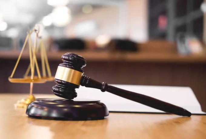 a wooden judge's gavel rests on its stand next to a golden scale of justice on a wooden desk, with a blurred courtroom…