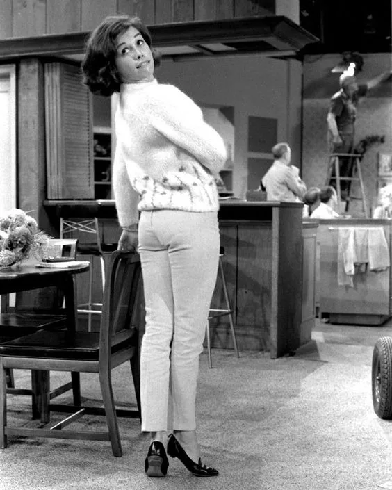 A black-and-white photo of a woman in a 1960s-style white fuzzy sweater and light pants, standing with her back to a chair…