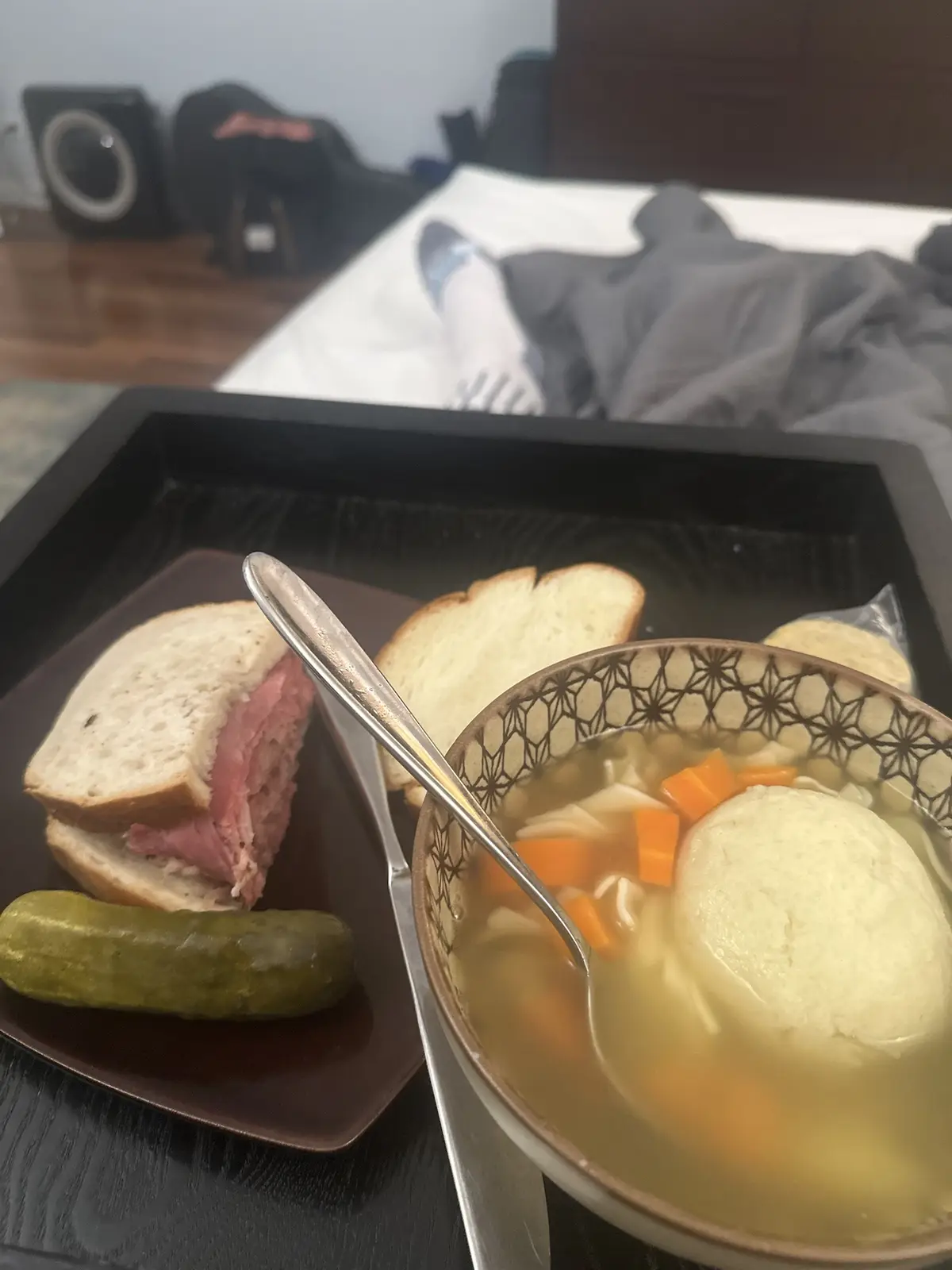 A black tray holds a meal consisting of a bowl of chicken noodle soup with a dumpling, a sandwich with deli meat, and a…