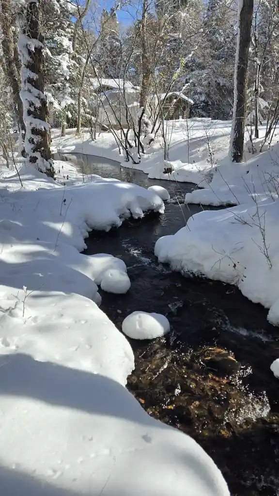 A serene winter scene shows a small stream flowing through deep snowdrifts, with snow-covered trees and a cabin visible in…