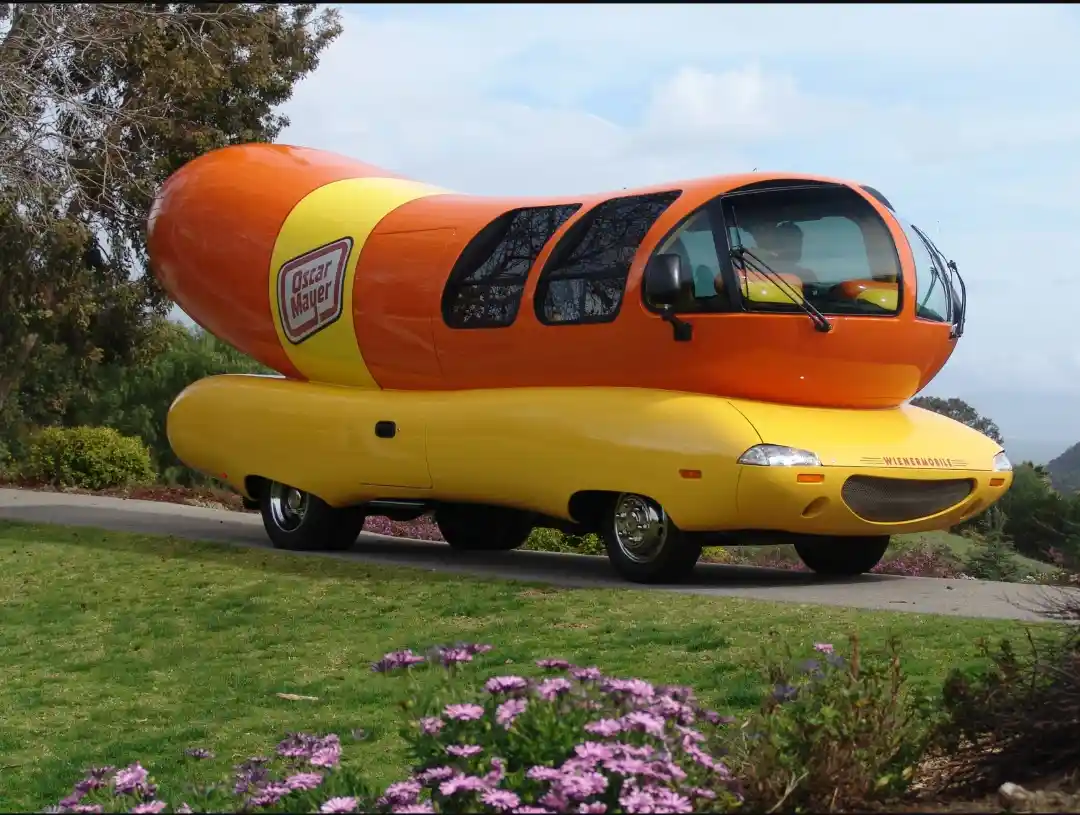 A large, orange-and-yellow vehicle shaped like a hot dog with the Oscar Mayer logo on its side, parked on a paved road…