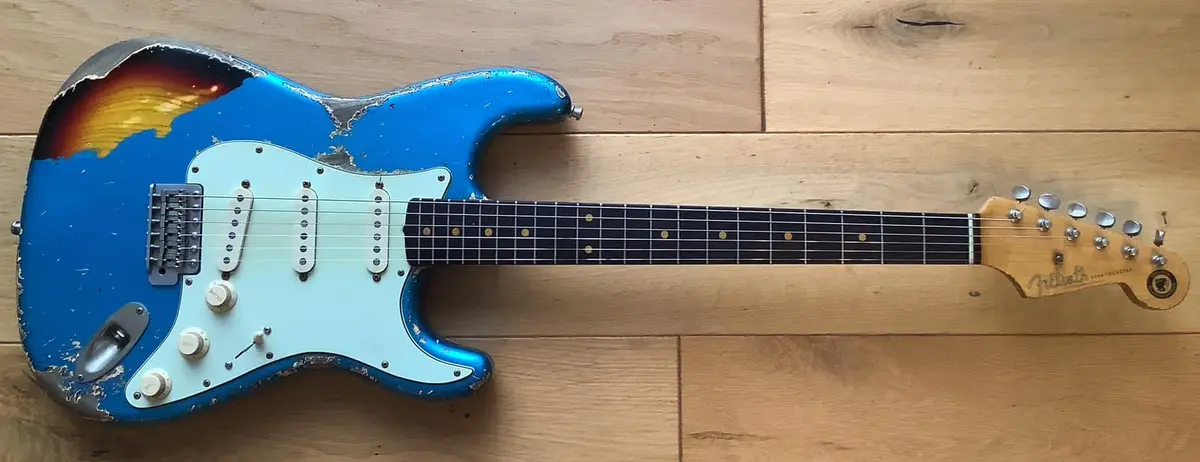 a heavily worn and distressed blue electric guitar with a sunburst finish lies on a wooden floor, its paint chipped and body…