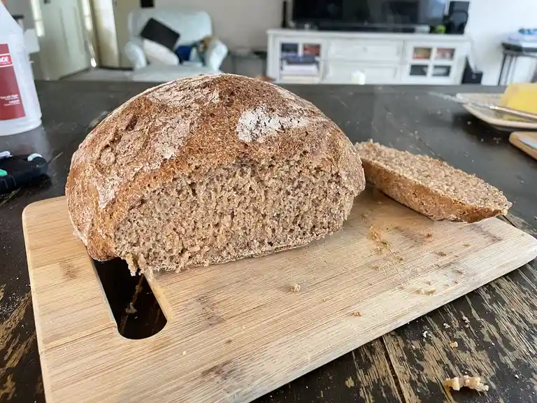 A rustic, round loaf of dark, whole-grain bread sits on a wooden cutting board with one slice cut and placed beside it. The…
