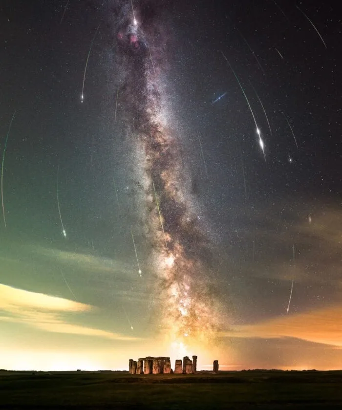 a long-exposure photograph shows the ancient stone circle of Stonehenge under a dramatic night sky. The Milky Way galaxy…