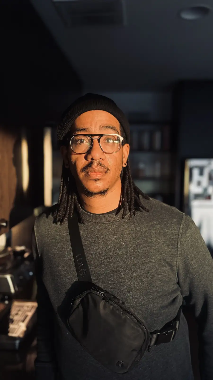A man with dreadlocks, glasses, and a black beanie stands in a dimly lit room with sunlight hitting his face. He wears a…
