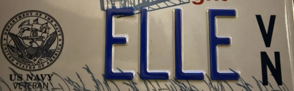 A close-up of a US Navy veteran license plate showing the name 'ELLE' in large blue letters, with 'V N' to the right. The…