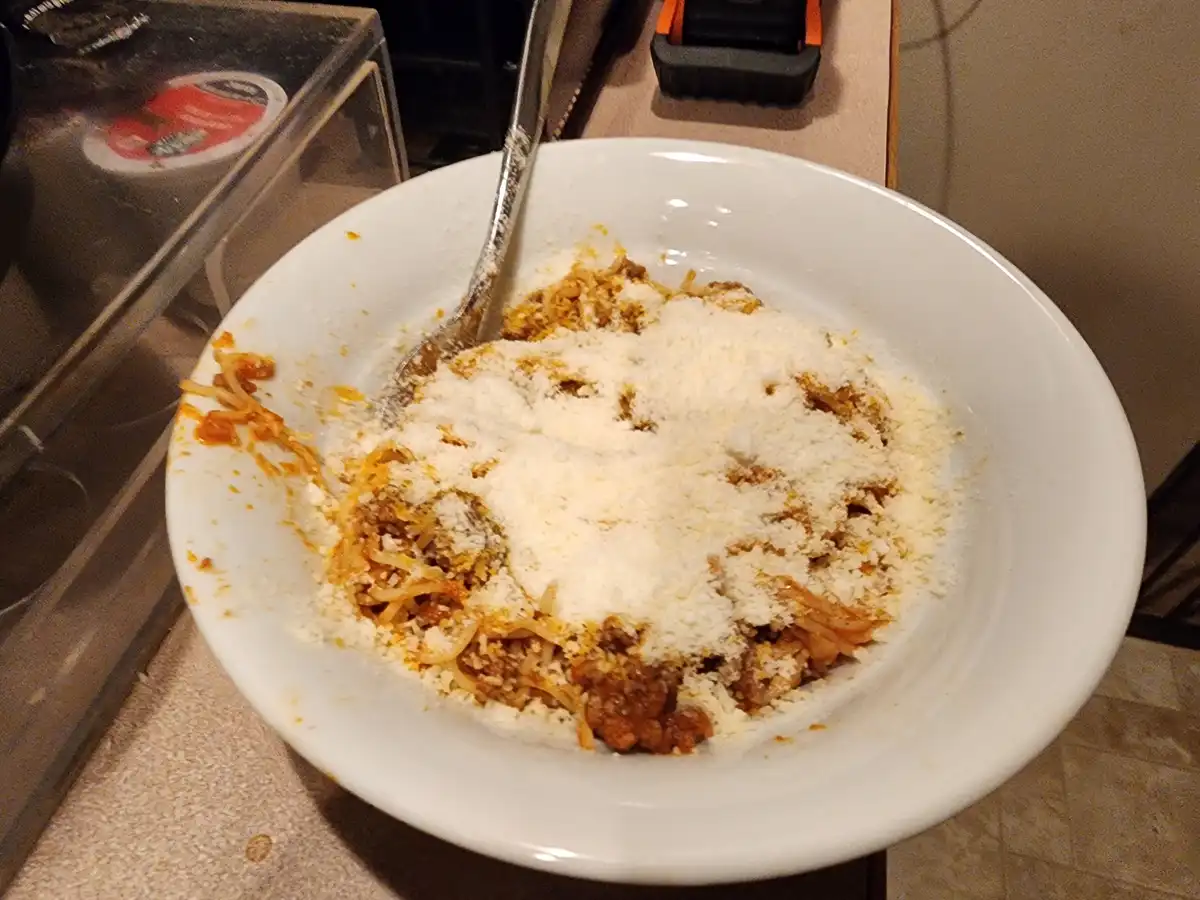 A white bowl of spaghetti with meat sauce and a heavy layer of grated parmesan cheese, mostly eaten with a fork resting in…