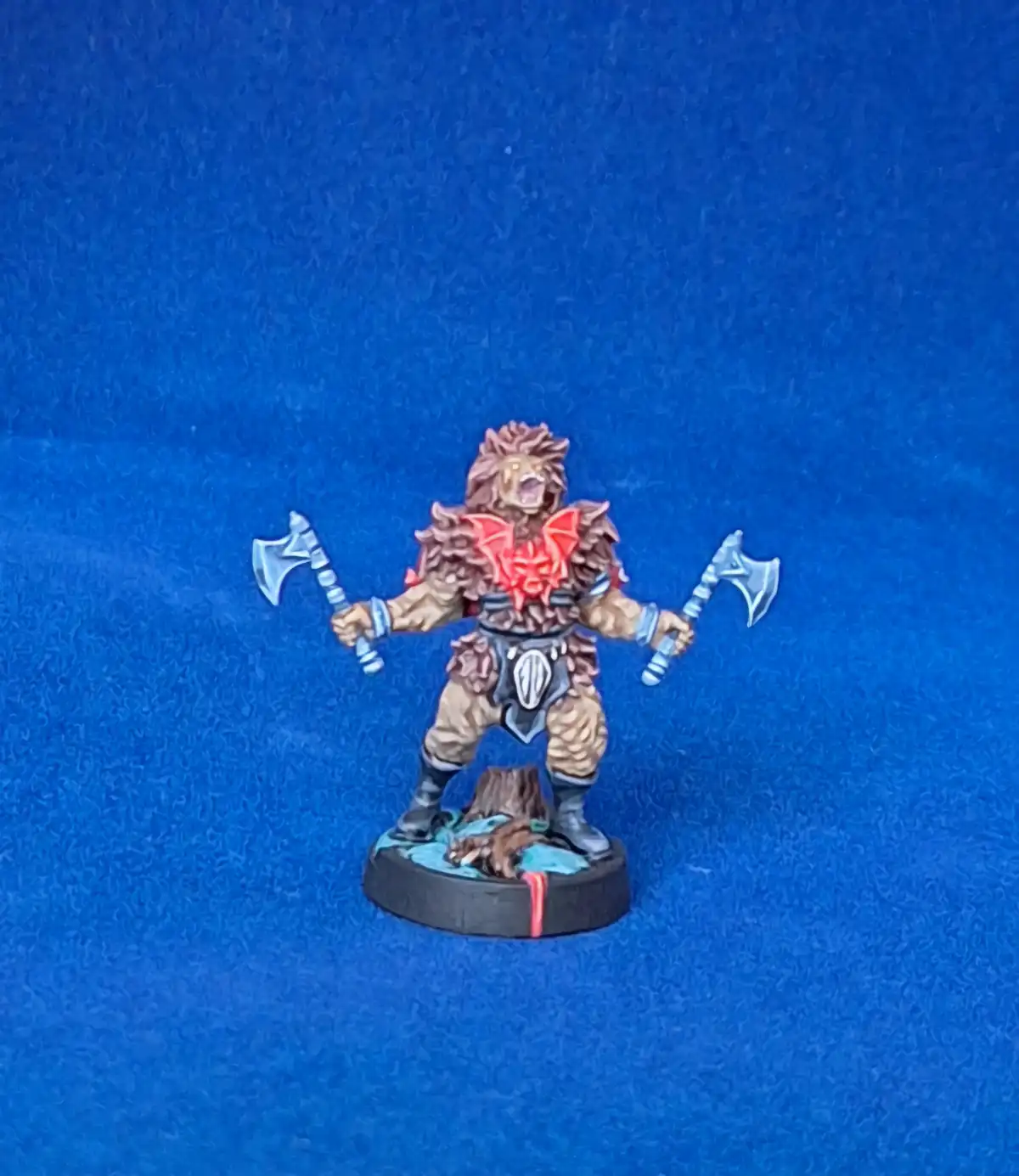 A painted miniature of a lion-headed humanoid warrior stands on a circular base against a solid blue background, holding one…