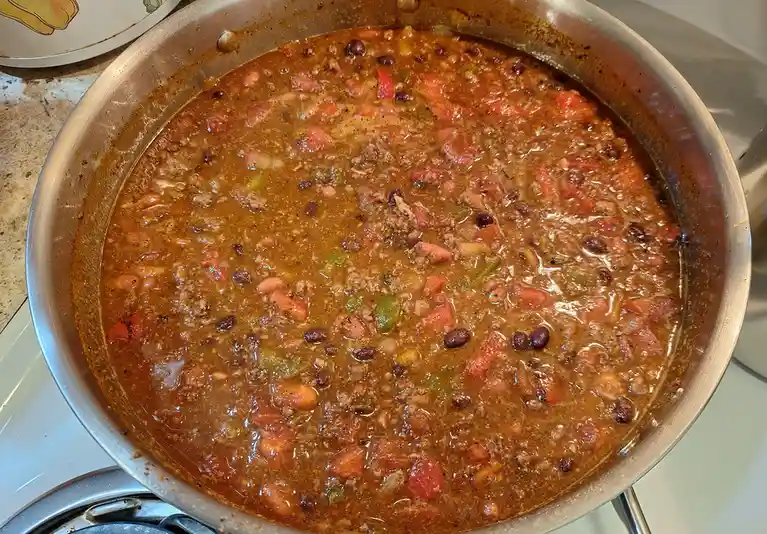 A close-up photo shows a large stainless steel pot filled with a thick, bubbling chili containing visible beans, ground…