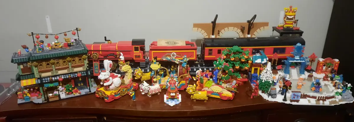 A wide shot of a wooden table displaying a large, elaborate Lego diorama featuring a red train set running through a festive…