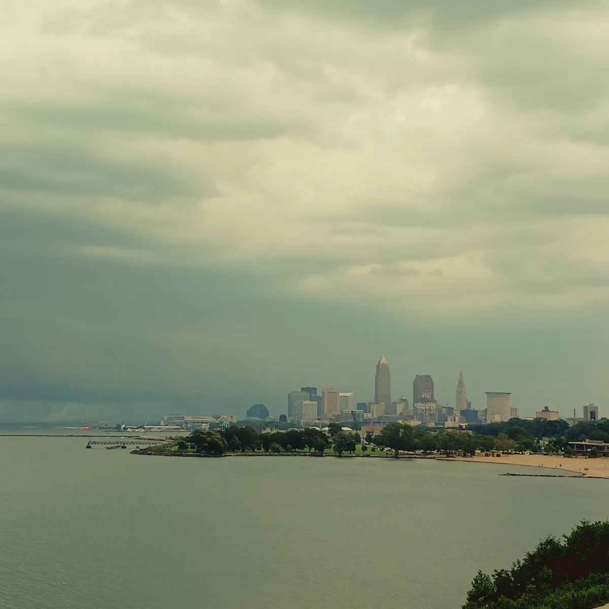 A wide shot of a city skyline, likely Cleveland, viewed across a body of water under a heavy, overcast sky. The foreground…