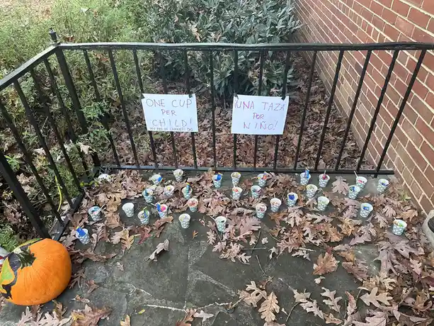 An outdoor Halloween scene shows dozens of small paper cups filled with candy arranged on a stone patio covered in fallen…