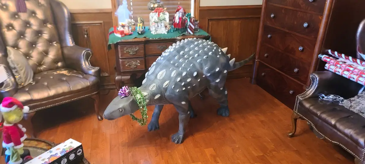 A large, grey Ankylosaurus dinosaur statue stands in a decorated living room, adorned with a green garland and a purple bow…