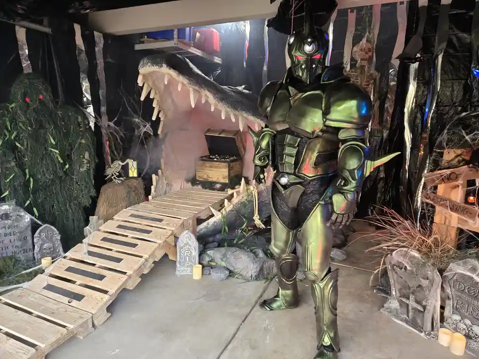 A person in a shiny green armored costume stands in a Halloween-themed setup with a large alligator head, tombstones, and…