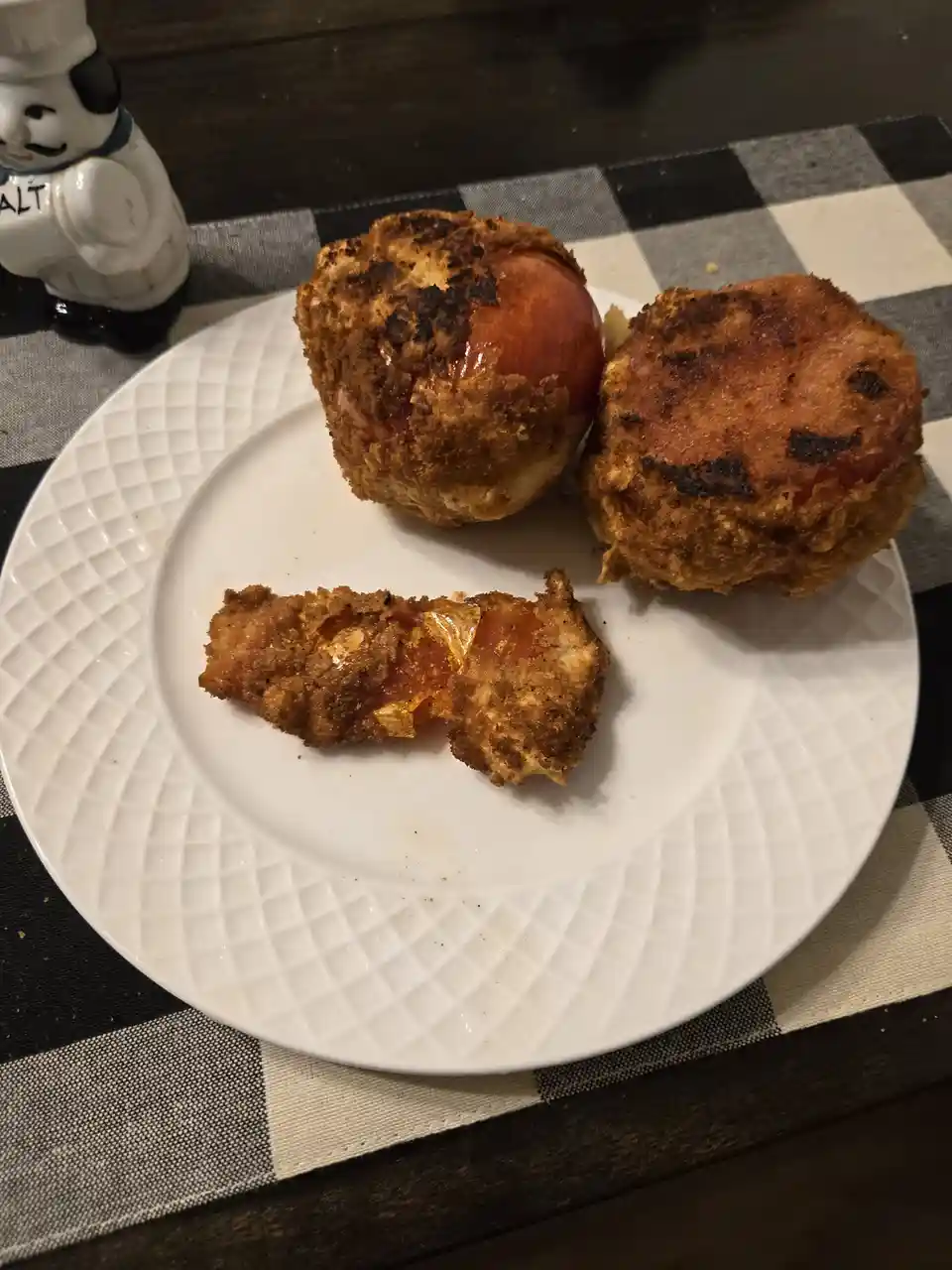 A white plate holds three fried tomatoes with crispy, burnt-looking breading; one is broken open to reveal the juicy red…