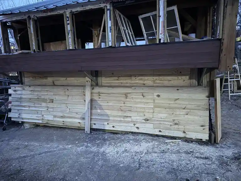 A partially constructed wooden house with a finished dark wood deck above and unfinished light wood siding below, showing…