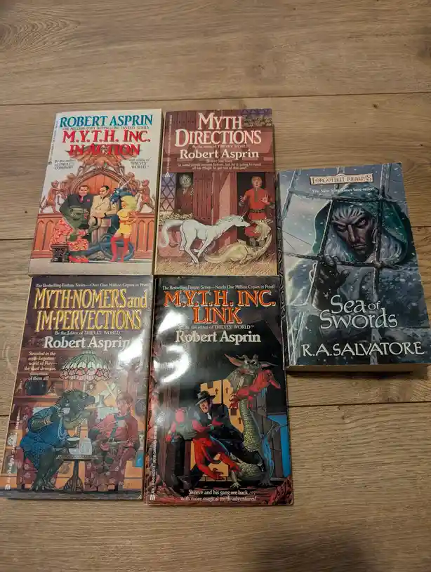 Five fantasy paperback books are arranged on a wooden surface. Four are by Robert Asprin, featuring titles like 'M.Y.T.H…