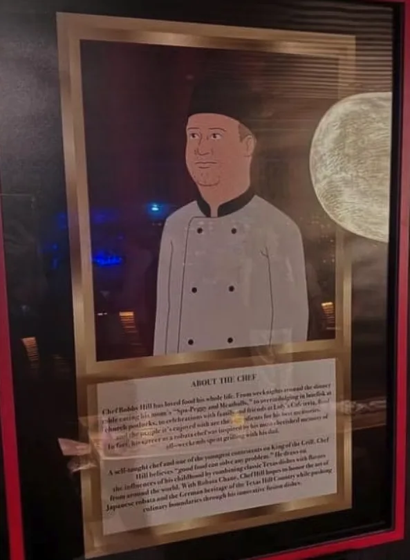 A framed portrait of a cartoon chef named Bobby Hill, with a printed bio below. The bio humorously claims he was inspired to…