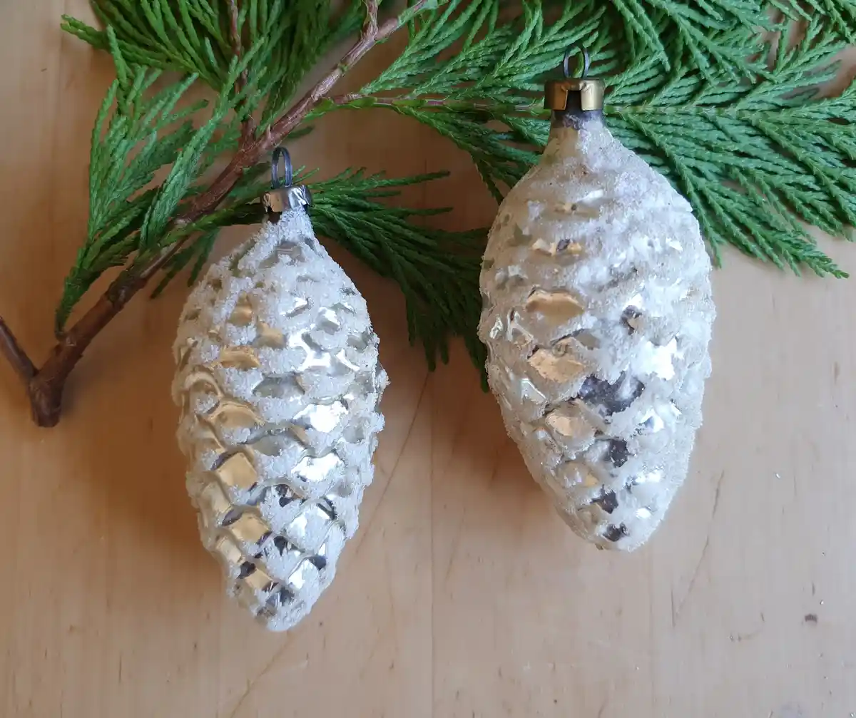 Two silver, pinecone-shaped glass Christmas ornaments covered in white sugar-like crystals rest on a wooden surface beside a…