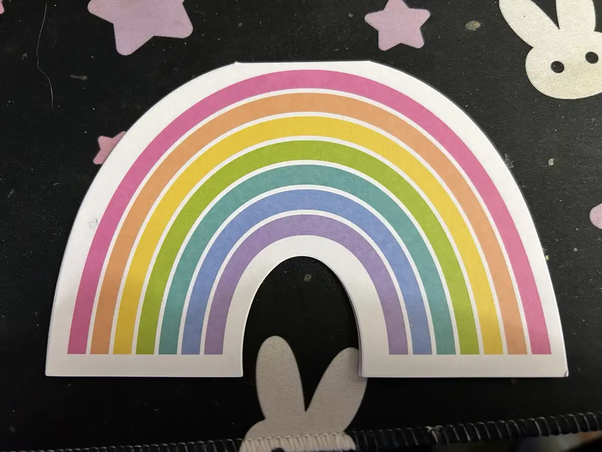 A colorful paper rainbow cutout with a white border is placed on a black surface decorated with pink stars and white bunny…