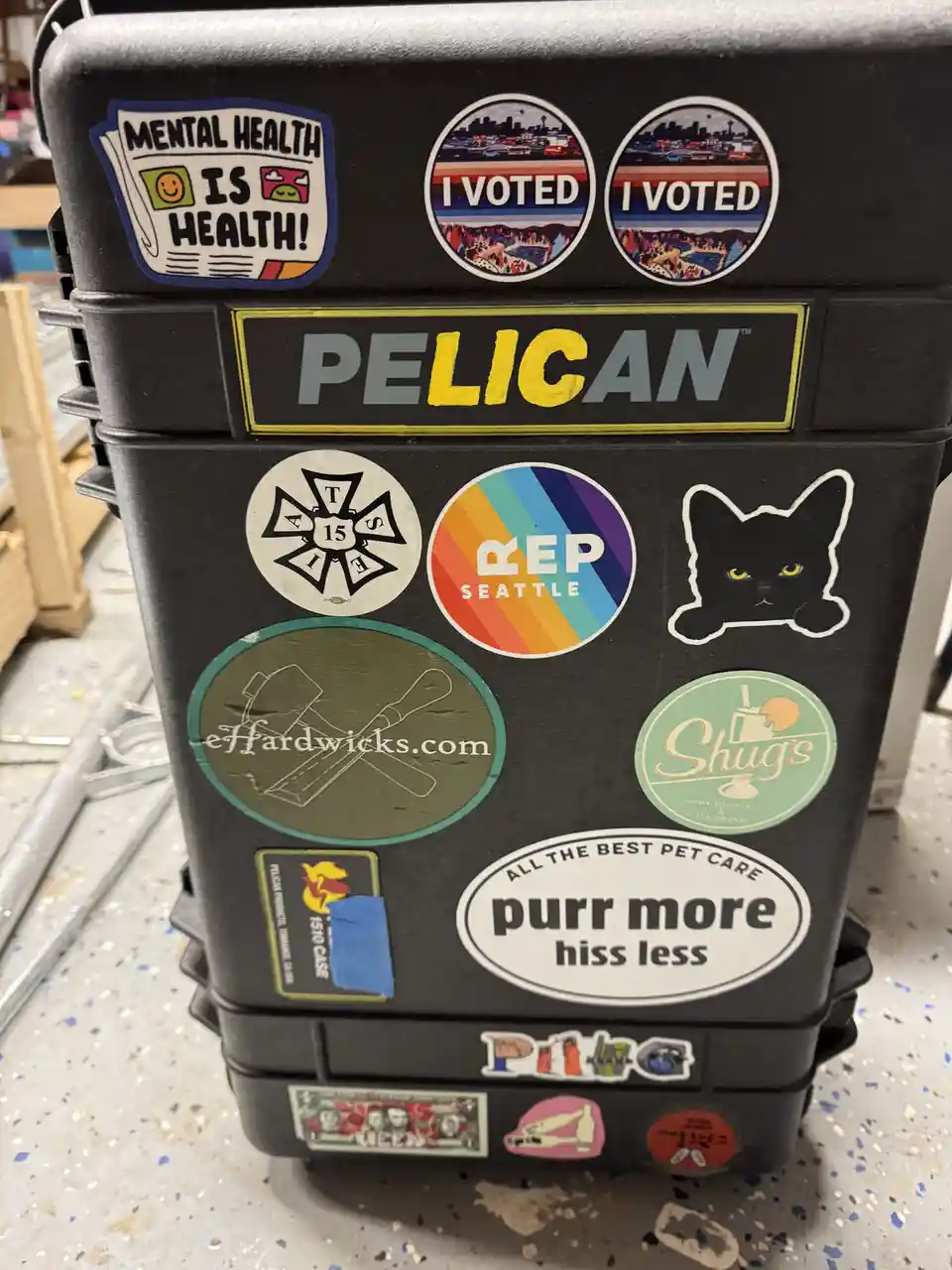 A black Pelican hard case is covered in numerous stickers, including 'Mental Health Is Health!', two 'I Voted' stickers, a…