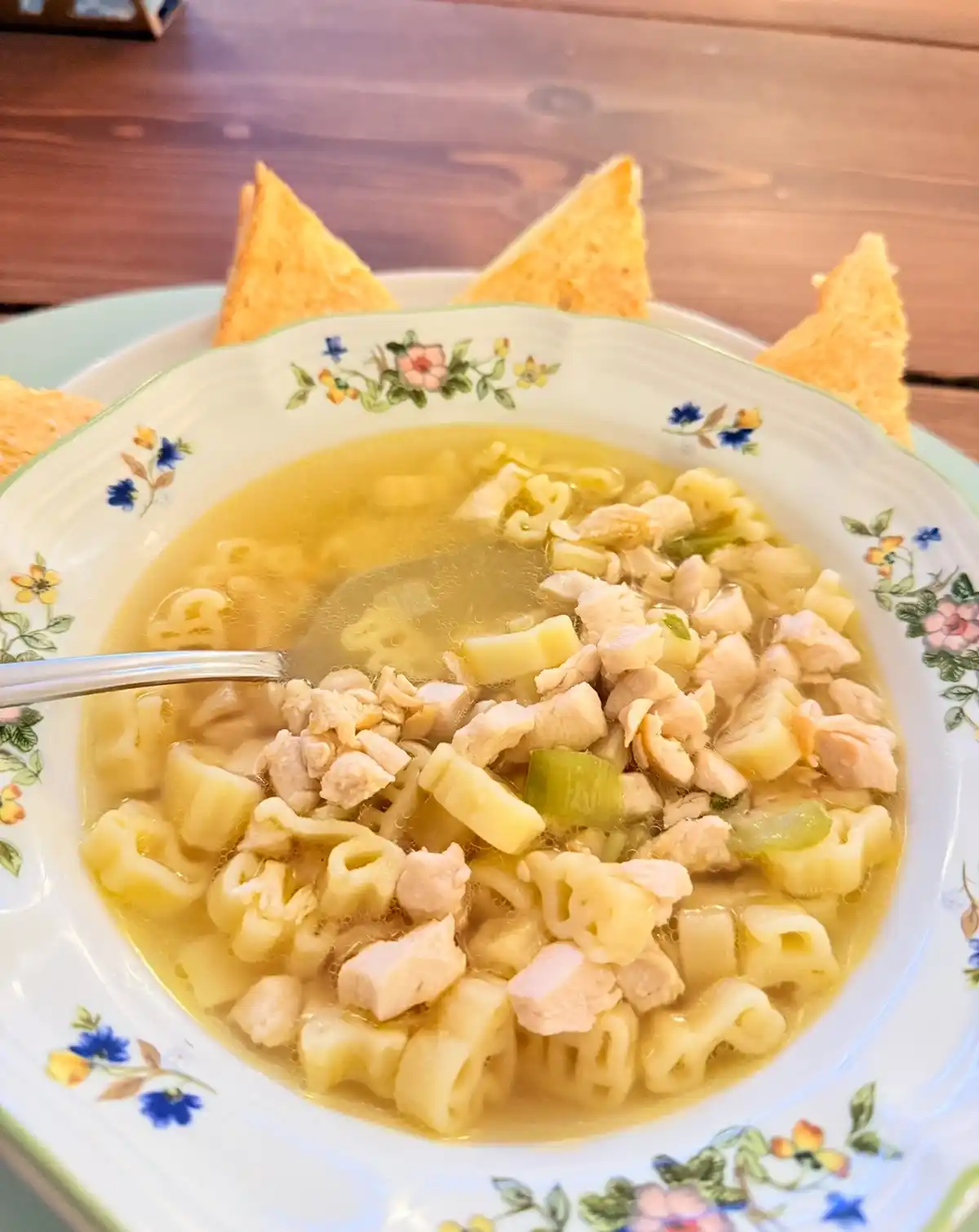 A close-up photo shows a bowl of chicken noodle soup with tortellini, chunks of chicken, and green vegetables in a clear…
