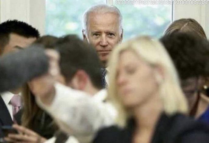 A man with white hair and a serious expression, identified as Joe Biden, is visible in the background of a crowded room…