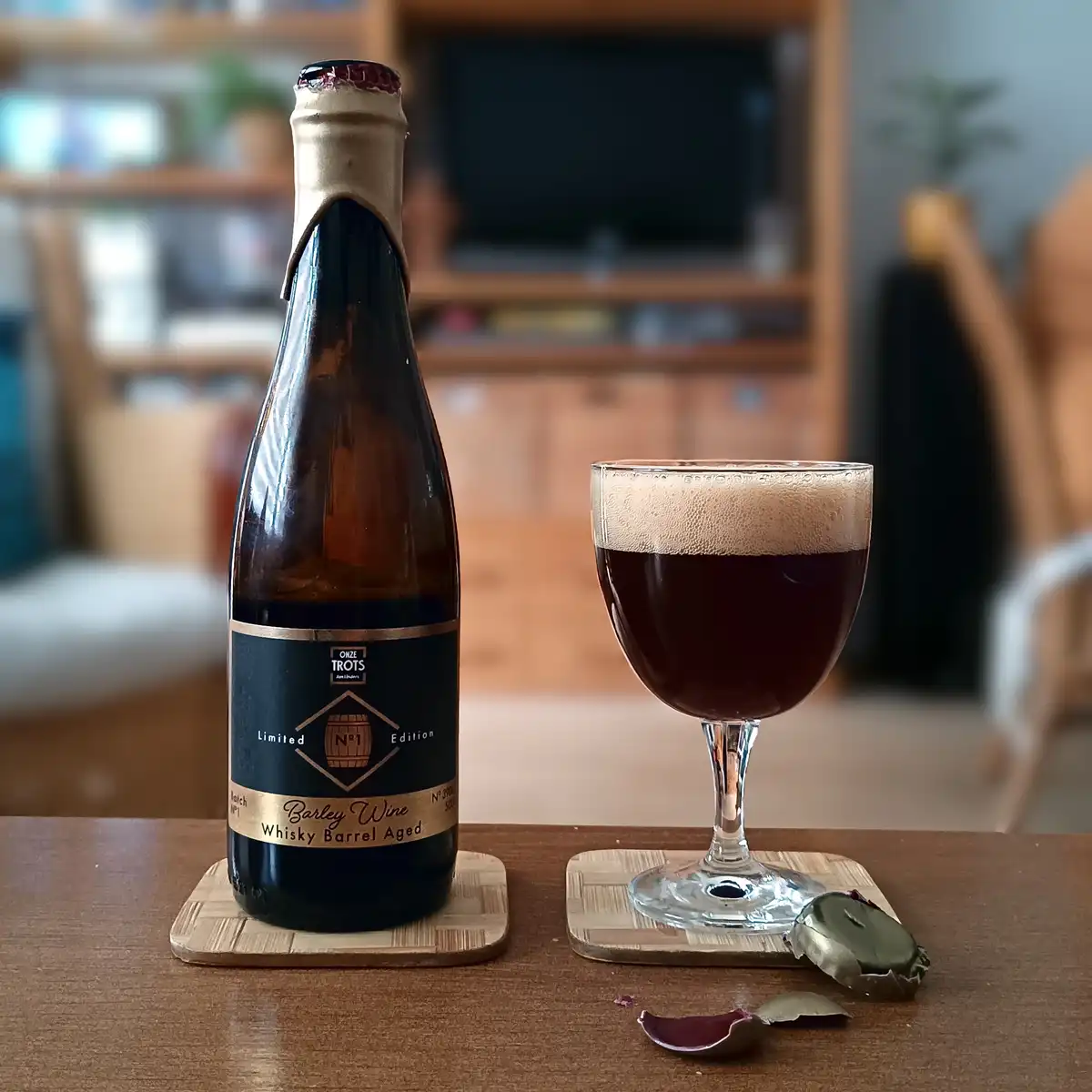 A bottle of 'Onze Trots' barley wine, labeled 'Limited Edition N°1 Whisky Barrel Aged', sits next to a glass filled with…