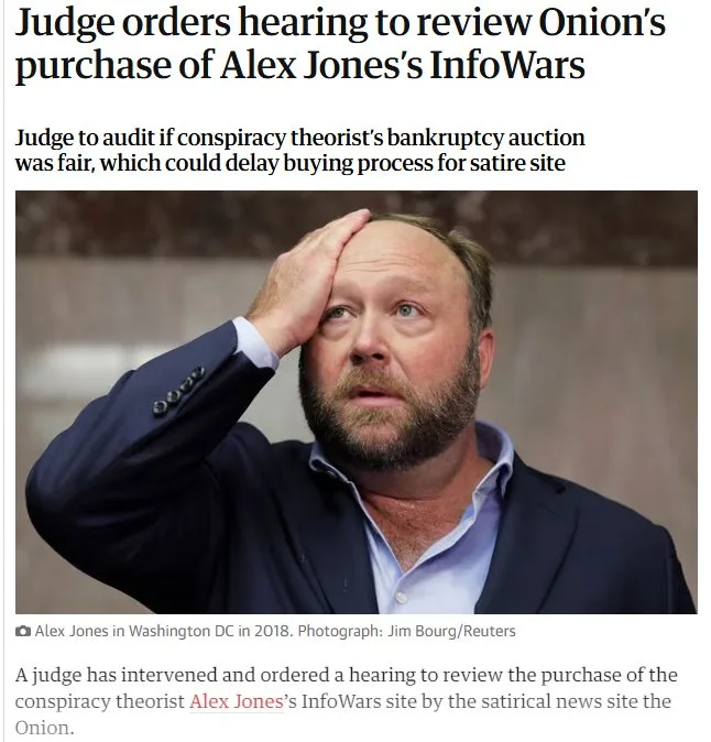 A man with a beard and blue eyes, wearing a dark suit jacket over a light blue shirt, holds his head in his hand with a…