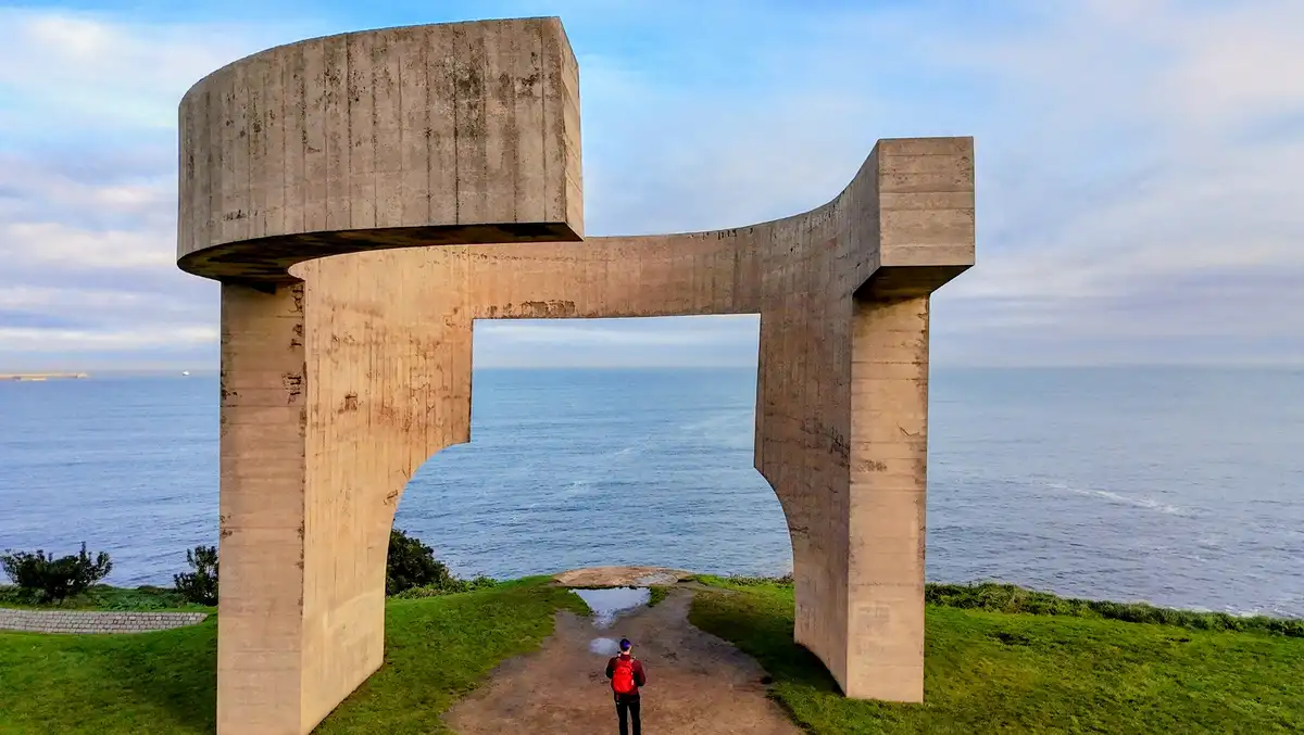 A person in a red backpack stands on a grassy path beneath a massive, curved concrete archway overlooking the ocean. The…