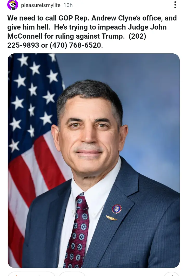 A formal headshot of a man in a dark suit and patterned tie, with an American flag visible behind him. The image has…