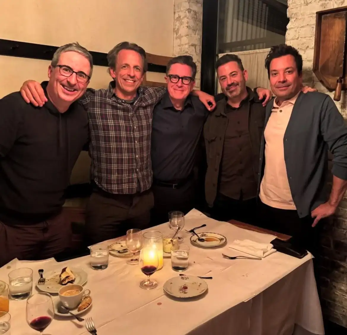 Five men are posing together at a restaurant table with empty plates and glasses, arms around each other, smiling for the…