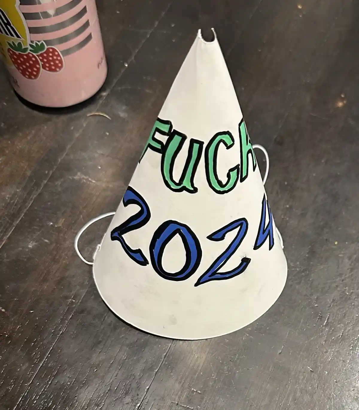 A white, cone-shaped party hat with hand-painted green and blue text sits on a dark wooden floor. The text reads 'FUCH' in…