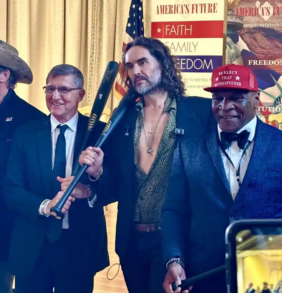 A man in a leopard-print shirt holds a baseball bat while standing between two men in suits at a political event. One man…