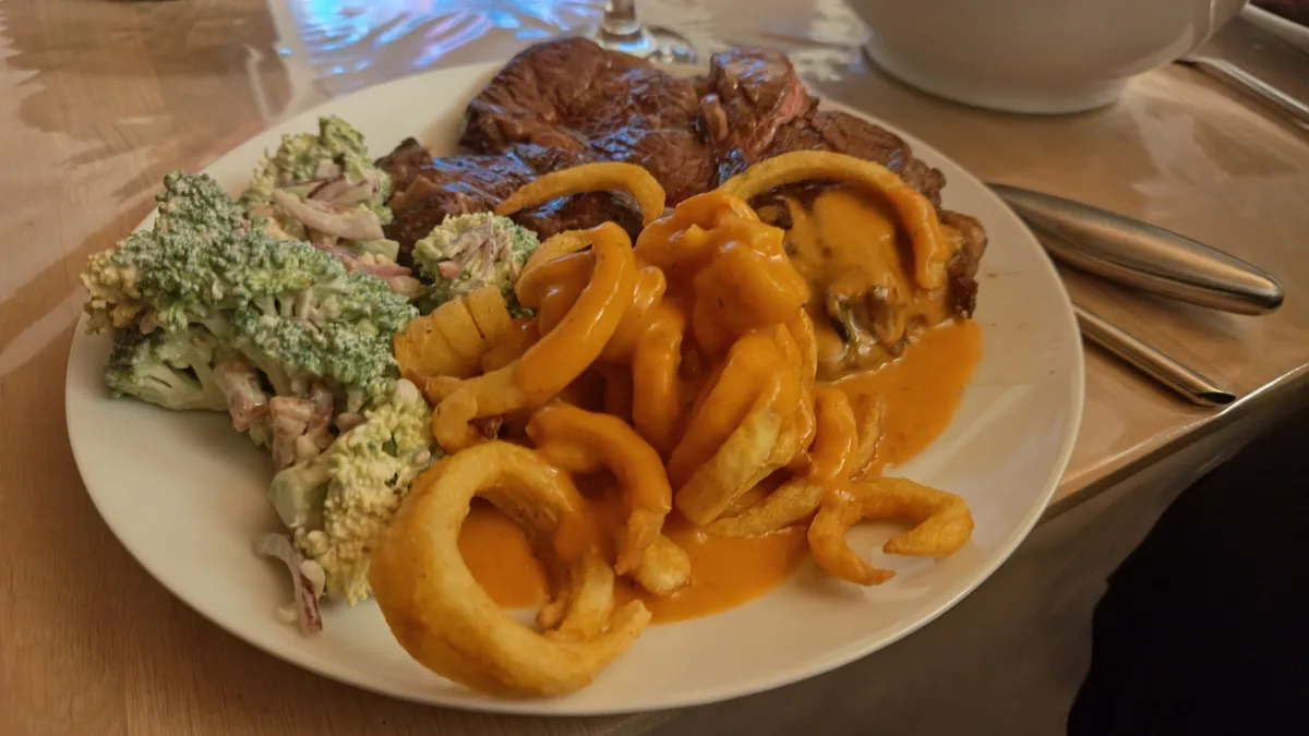 A plate of food features a large, seared steak, a side of broccoli salad, and curly fries covered in orange cheese sauce…