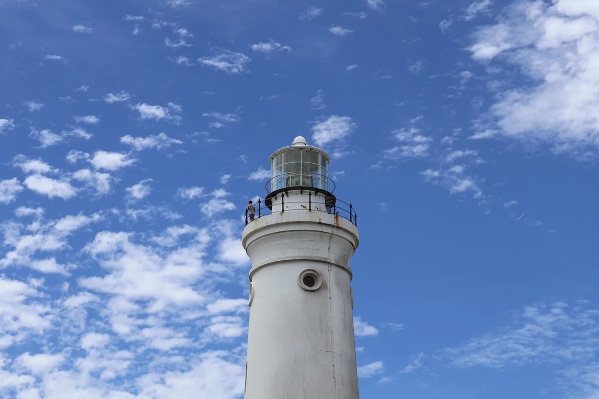 A white lighthouse stands against a bright blue sky with scattered white clouds. Two people are visible on the railing at…