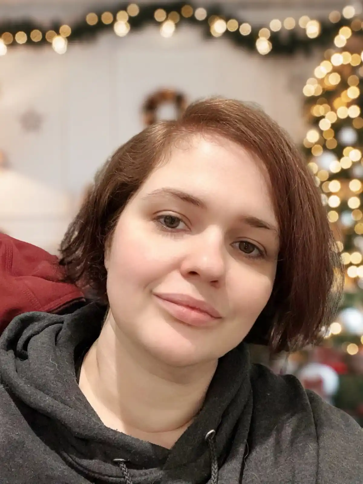 A selfie of a woman with short brown hair wearing a dark grey hoodie, smiling slightly with a hint of sadness or…
