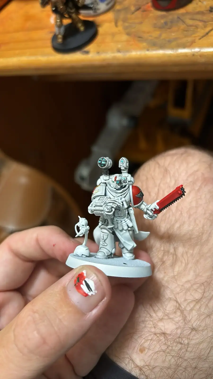 A hand holds a small, unpainted white miniature of a sci-fi armored warrior holding a red chainsaw. The thumb nail has a…