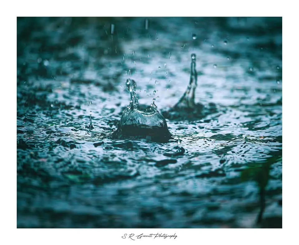 A high-resolution photograph captures raindrops splashing into a body of water, creating crown-like ripples and droplets…