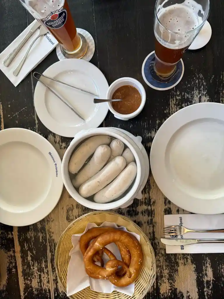 An overhead shot of a dark wooden table set with German-style food: a white ceramic pot filled with raw, uncooked sausages…
