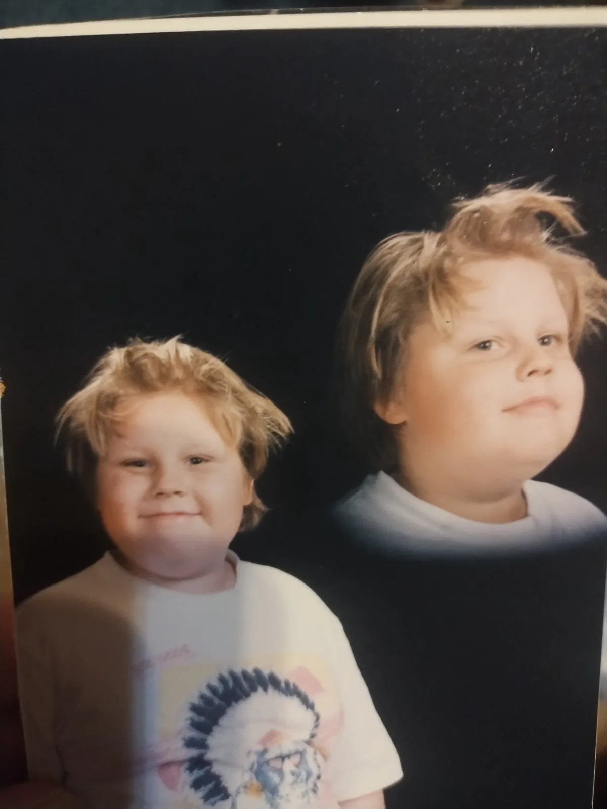 A vintage childhood photo shows two young boys with similar blond hair and chubby faces against a dark background. The boy…