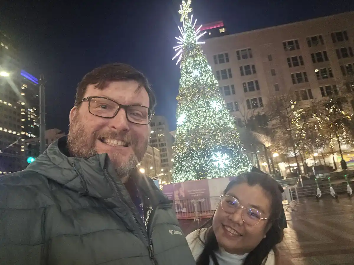 A selfie of a smiling man and woman at night in front of a large, brightly lit Christmas tree in a city plaza. The man wears…