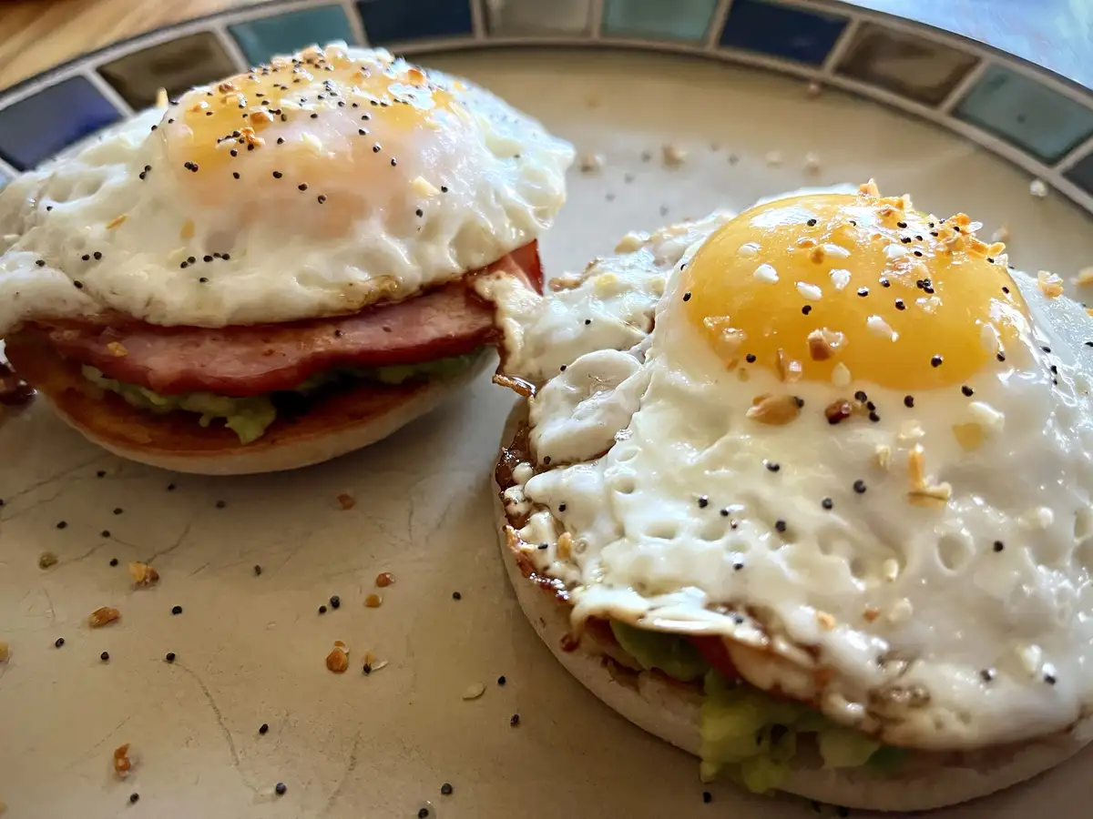A close-up photo shows two breakfast sandwiches on English muffins, each topped with a fried egg, avocado, and ham. The egg…