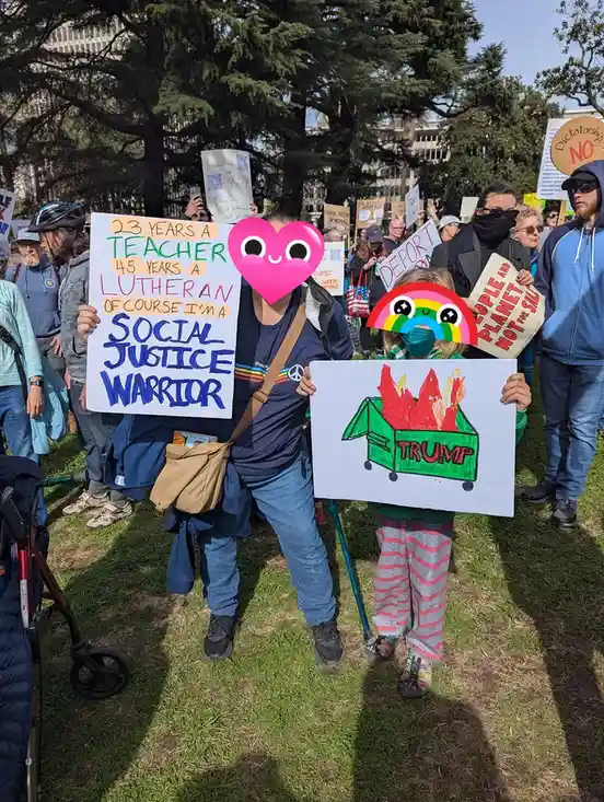 A person at a protest holds a handmade sign stating '23 YEARS A TEACHER 45 YEARS A LUTHERAN OF COURSE I'M A SOCIAL JUSTICE…