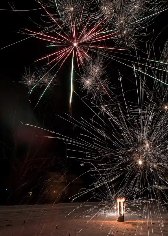 A long-exposure photograph captures multiple fireworks exploding in the night sky above a snowy ground, with a single…