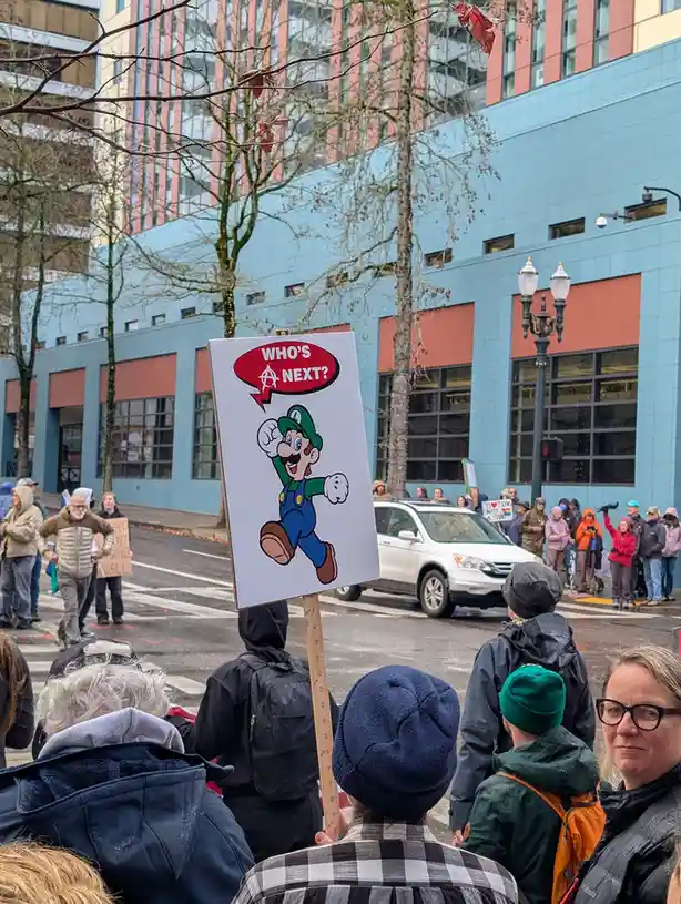 A protest scene with a crowd of people outdoors, one holding a sign featuring the video game character Luigi with a red…