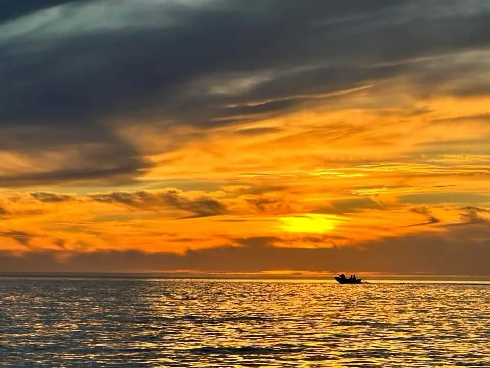 A serene sunset over the ocean with dark clouds above and golden-orange light reflecting on the water. A small boat with a…