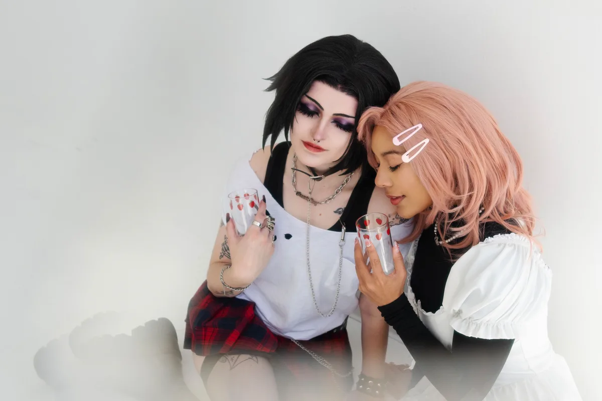 Two people in cosplay, one with black hair and gothic makeup holding a glass with strawberry decals, the other with pink…